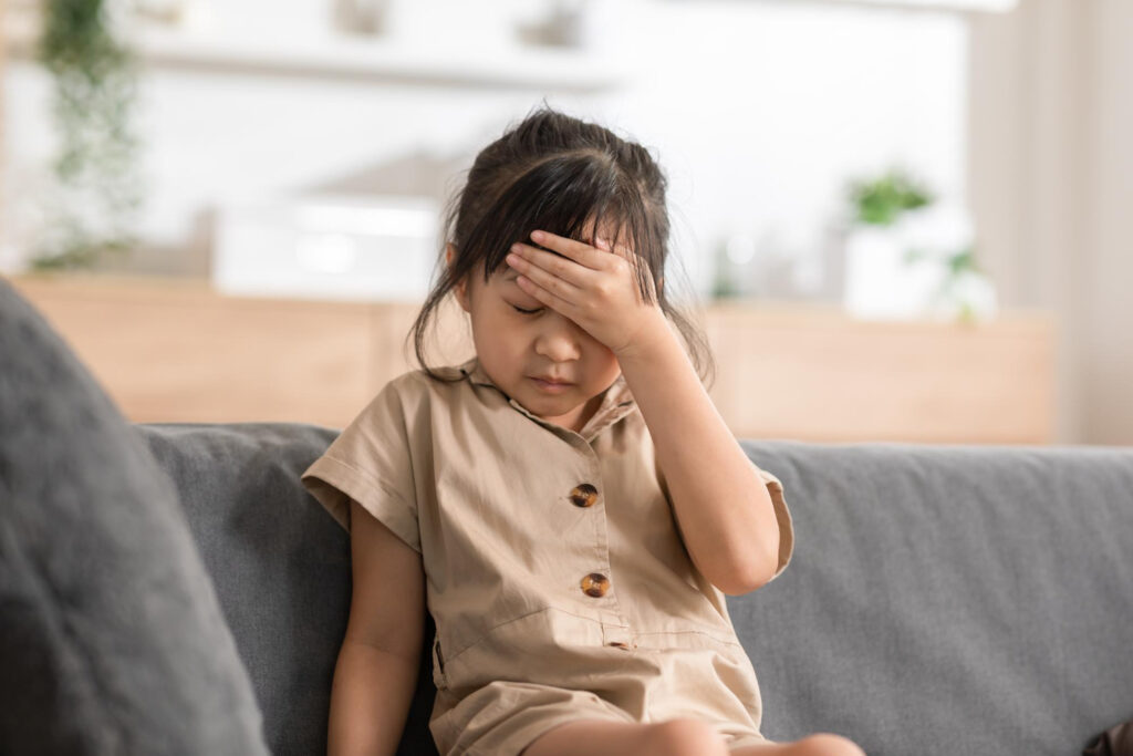 Young girl sits on a gray sofa, hand pressed to her forehead in a tired or stressed gesture.