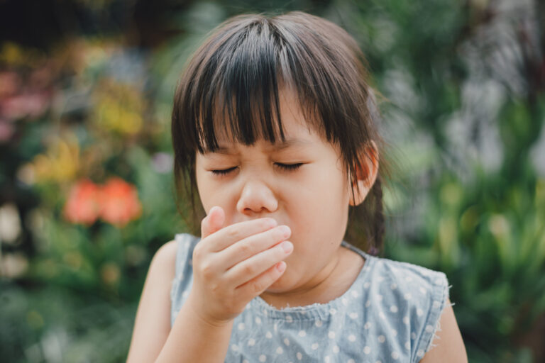 Young girl in a light blue polka-dot dress covers her mouth with her hand outdoors, appearing to cough or yawn in a garden.